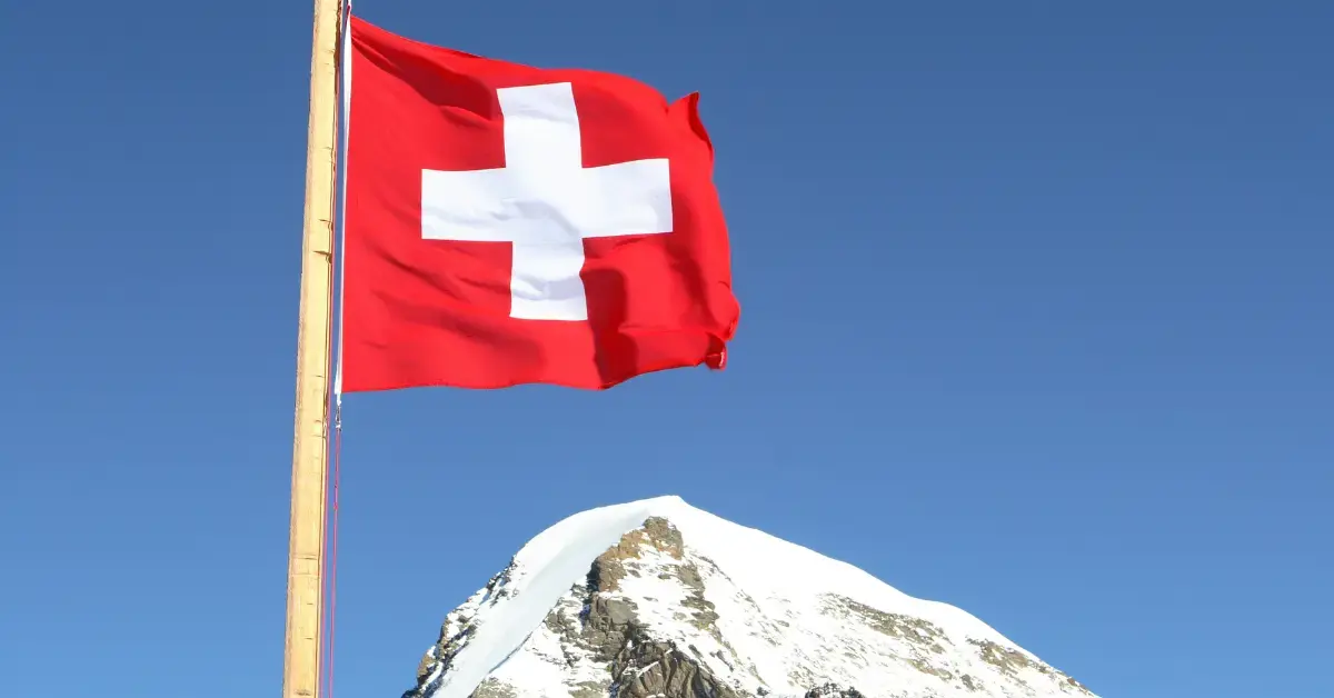 Schweizer Flagge auf einem Holzmasten weht vor einem verschneiten Alpengipfel unter blauem Himmel